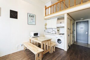 A compact kitchen with a microwave, sink, and washing machine. There's a wooden table and bench, and a staircase leading to an upper level.