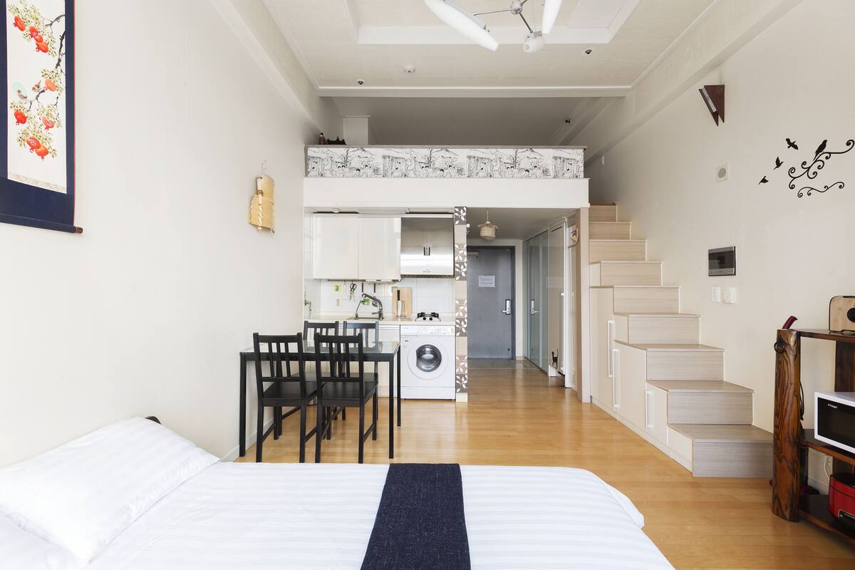 Habitación de hotel pequeña con cama, escritorio con sillas, lavadora y una escalera que lleva a un nivel superior.