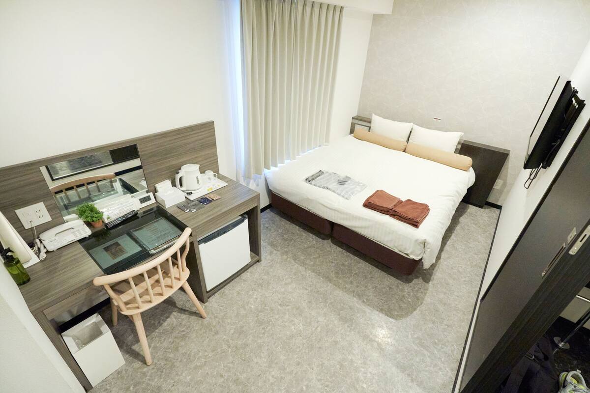 Quarto de hotel com cama, escrivaninha com computador, cadeira, mesinha e televisão.
