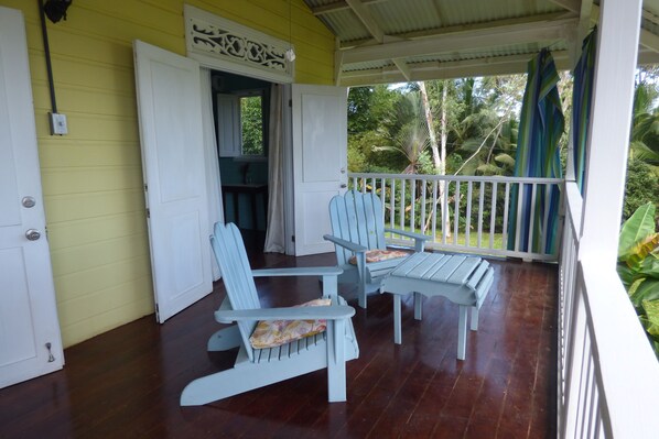 A porch with two Adirondack chairs and a bench, a white door, and a view of greenery.
