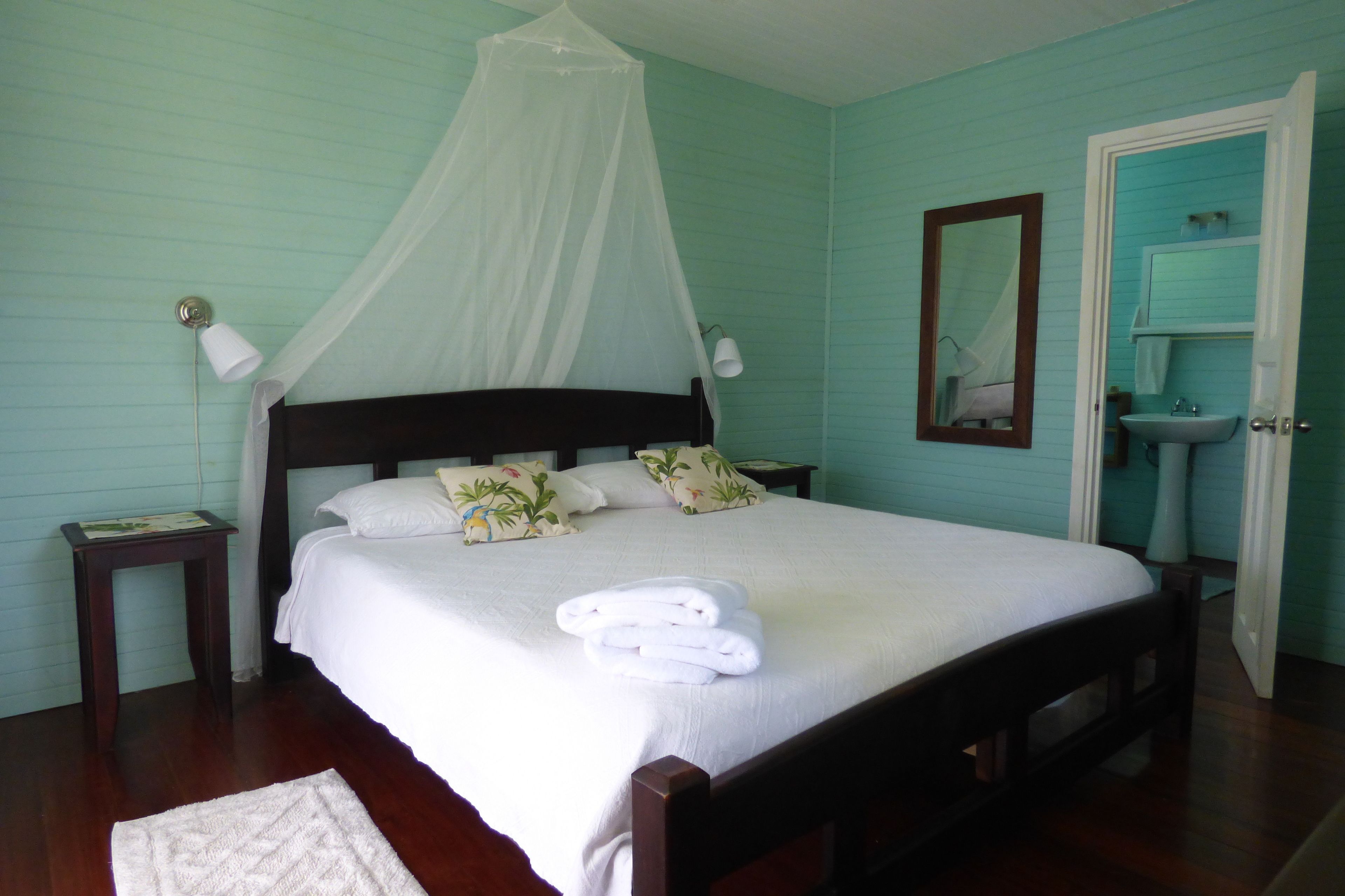 A neatly made bed with white linens and a canopy, a bedside table with a lamp, and a bathroom with a sink and mirror.