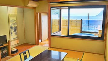 A traditional Japanese room with tatami flooring, a tatami mat table, a low wooden table, a television, and a view of the sea through a large window.