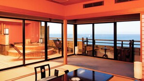 Una habitación con un amplio ventanal corredera que da a un balcón con vista al océano. En el interior, hay una mesa de madera con sillas, un pequeño rincón de estar con un banco y piso de madera.
