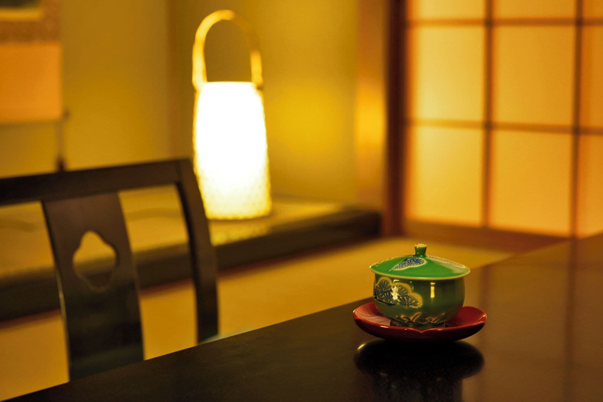 Een tafel met een groene kom op een rode schotel, een zwarte stoel en een brandende lamp op de achtergrond.