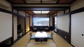 Una habitación japonesa tradicional con suelo de tatami, una mesa baja con cojines, un televisor y vistas a las montañas a través de un ventanal grande.