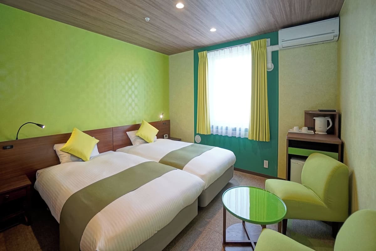 A room with two beds, a small table, a chair, and a kitchenette.