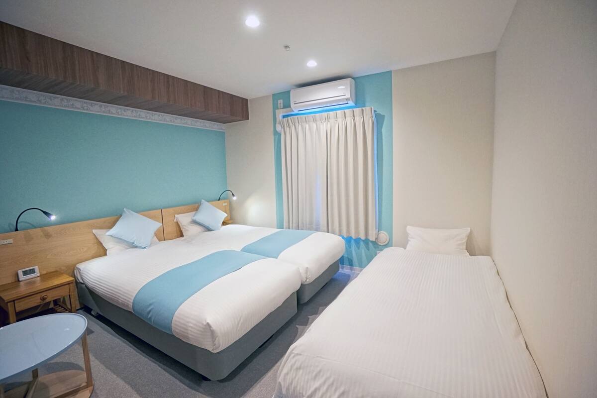 A hotel room with two beds, a bedside table, a chair, an air conditioning unit, and a window with curtains.