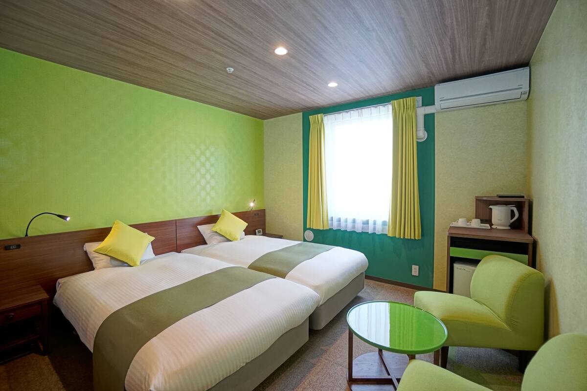 A hotel room with two beds, a small table, a chair, and a window with curtains.