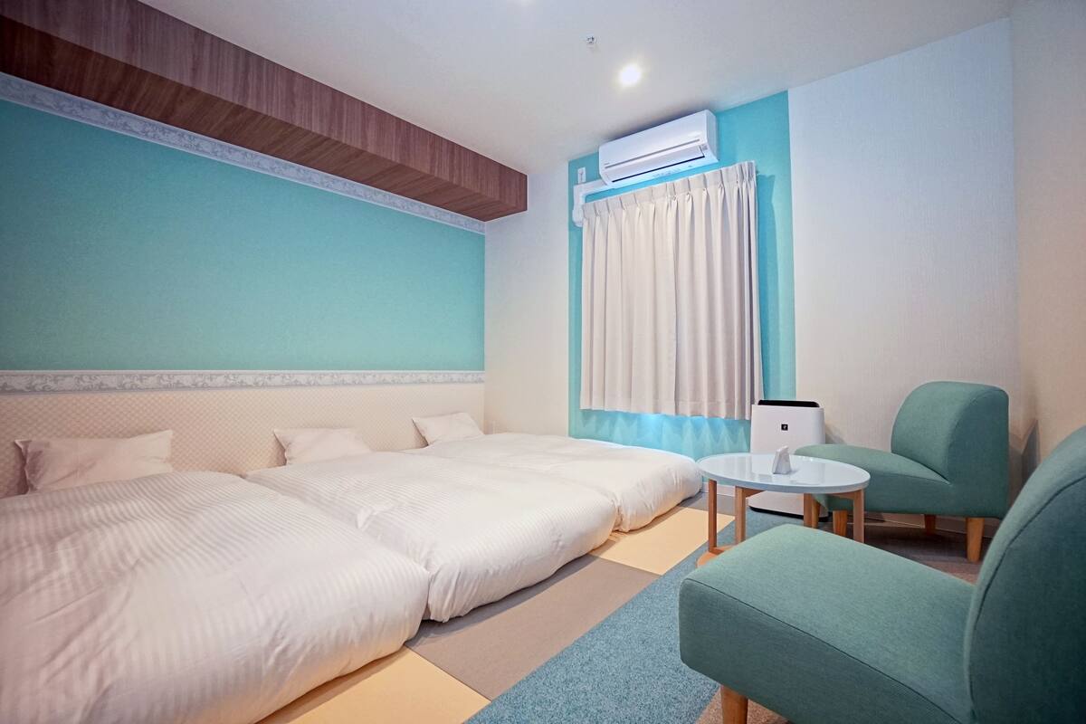A hotel room with two beds, a small round table, a green armchair, and an air conditioning unit.