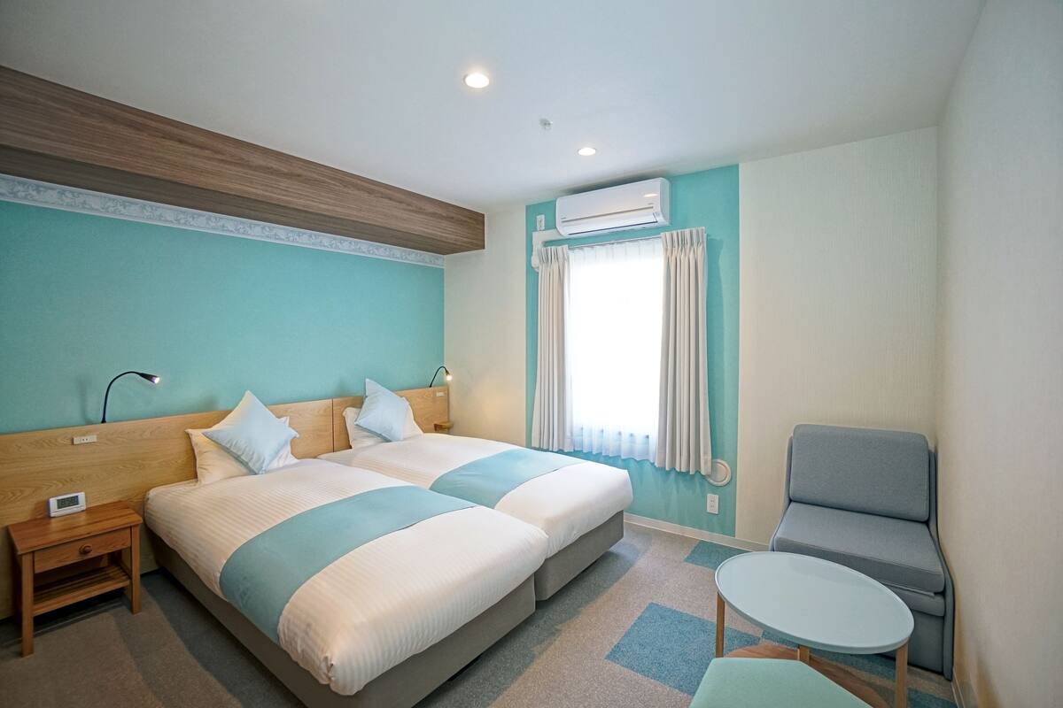 A hotel room with two beds, a small table, a chair, an air conditioning unit, and a nightstand with a lamp.