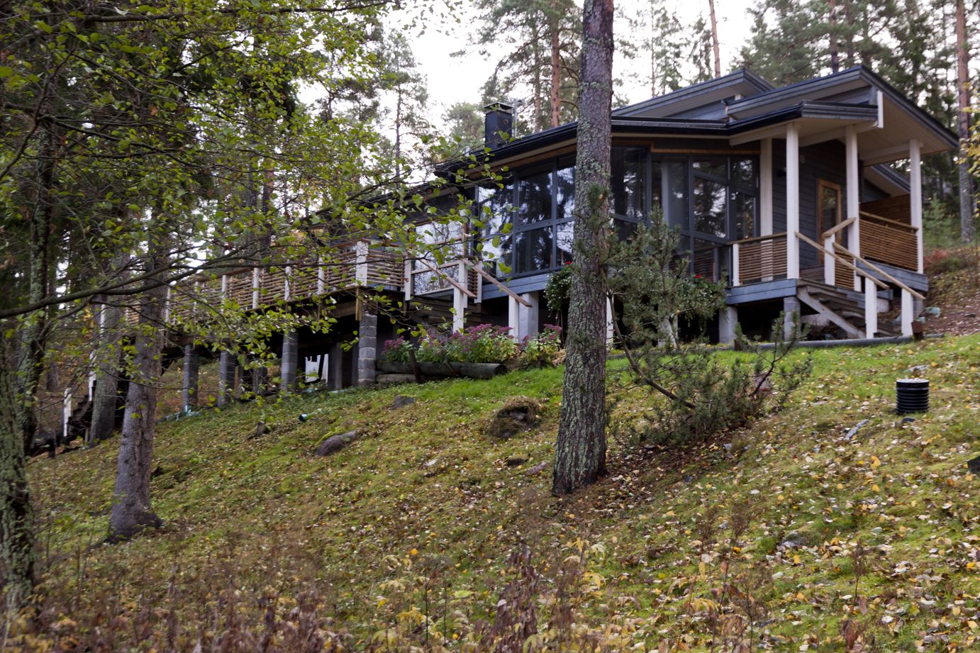 A modern cabin with a wooden deck and a glass facade, surrounded by a forested hillside.