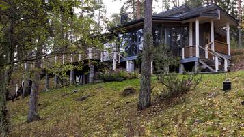 A modern cabin with a wooden deck and a glass facade, surrounded by a forested hillside.