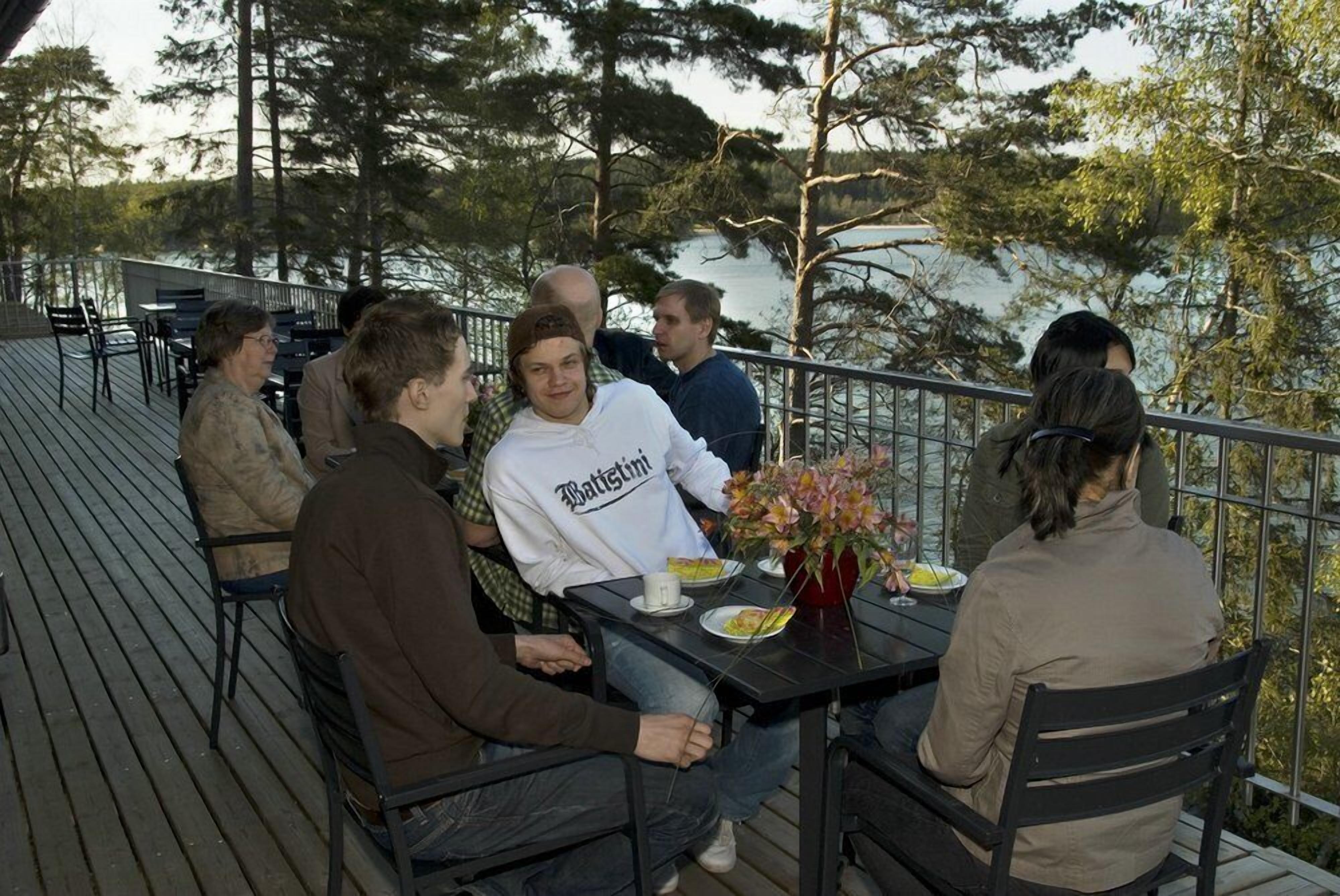 Een groep mensen zit aan tafels op een terras met uitzicht op bomen en water.
