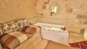 A room with a stone wall, a white bathtub with rose petals, a patterned sofa, and a small mirror.