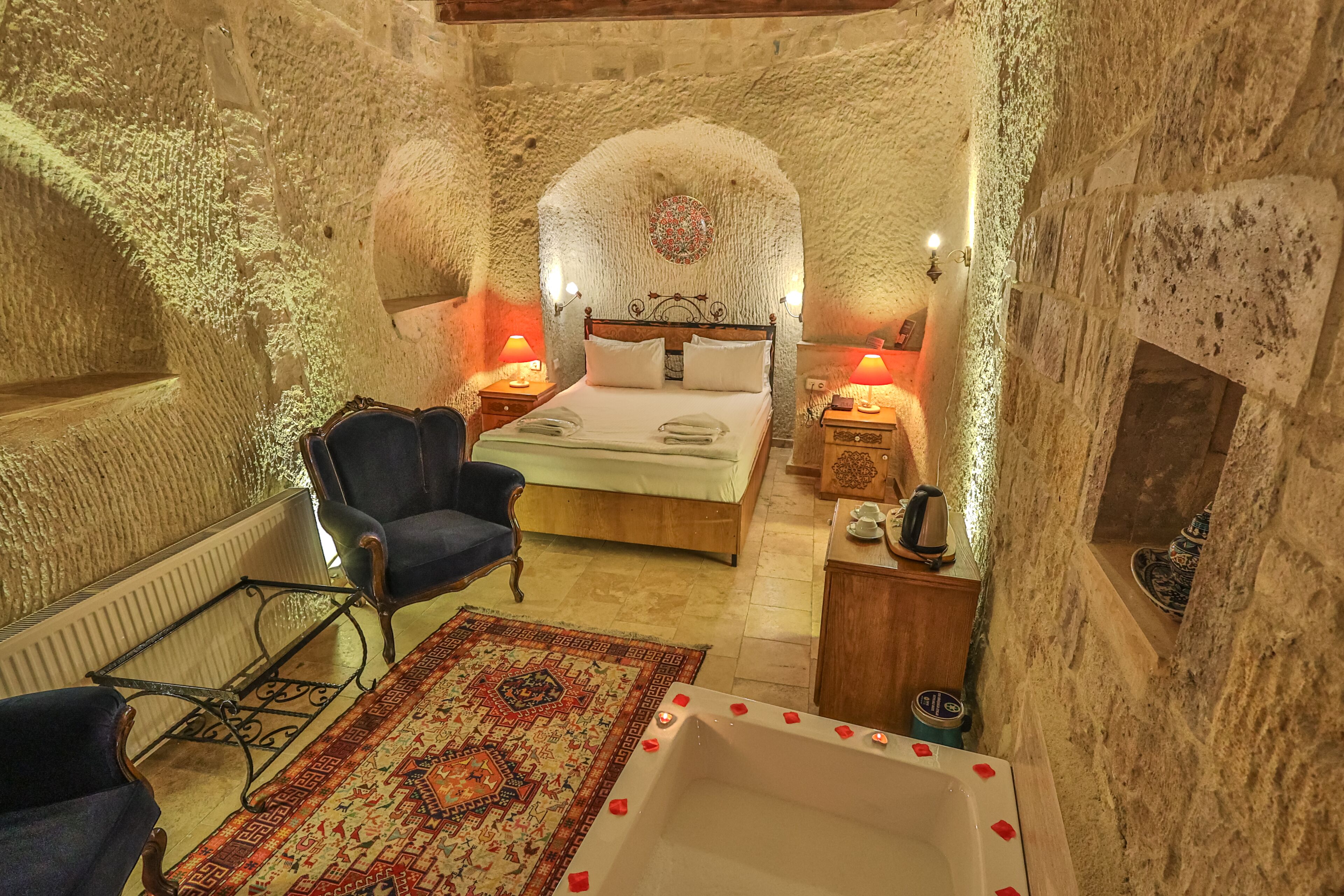 A stone-walled bedroom with a bed, two bedside tables, a chair, a rug, and a bathtub.