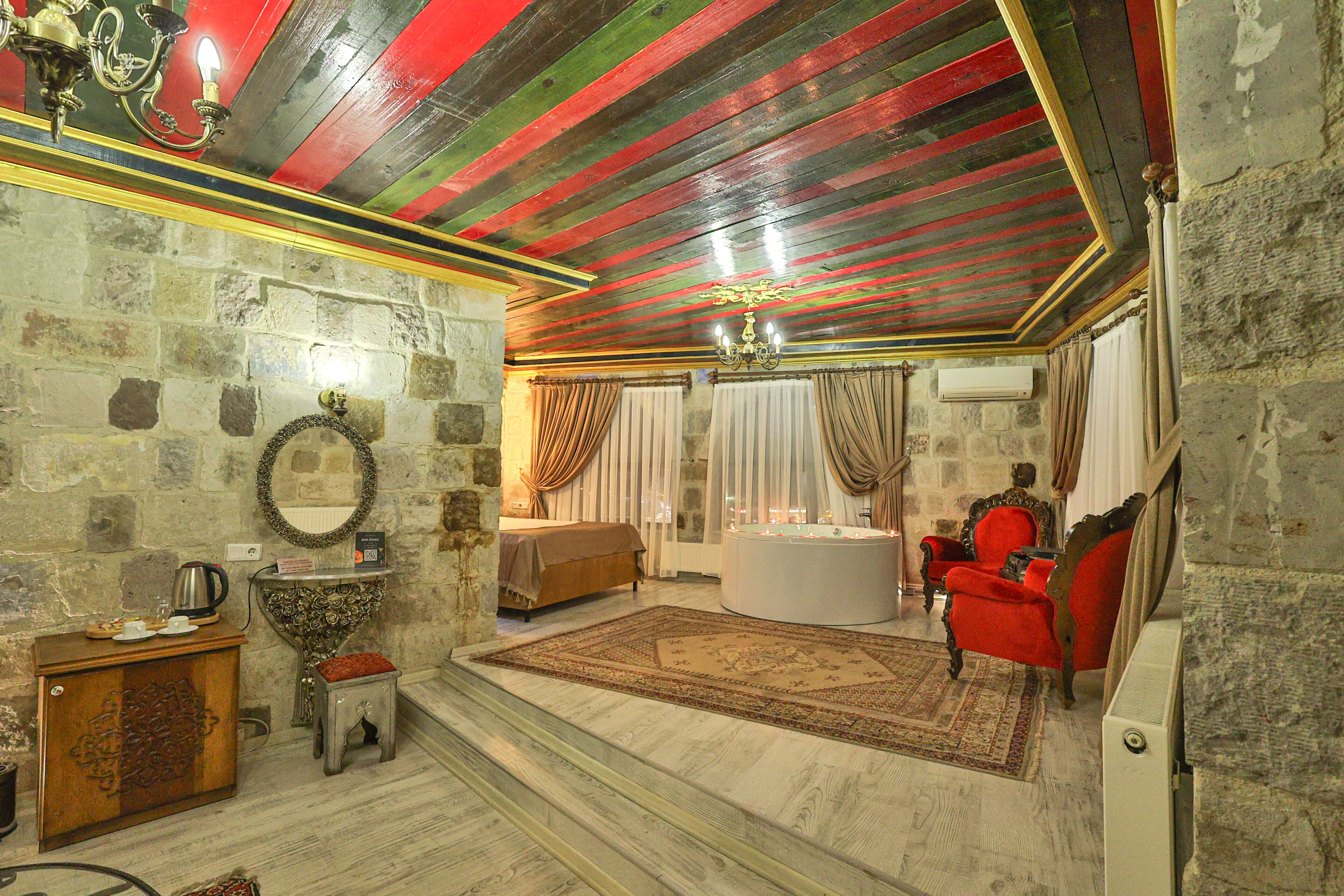 A room with a stone wall, a wooden cabinet, a red armchair, a white bathtub, and a chandelier.