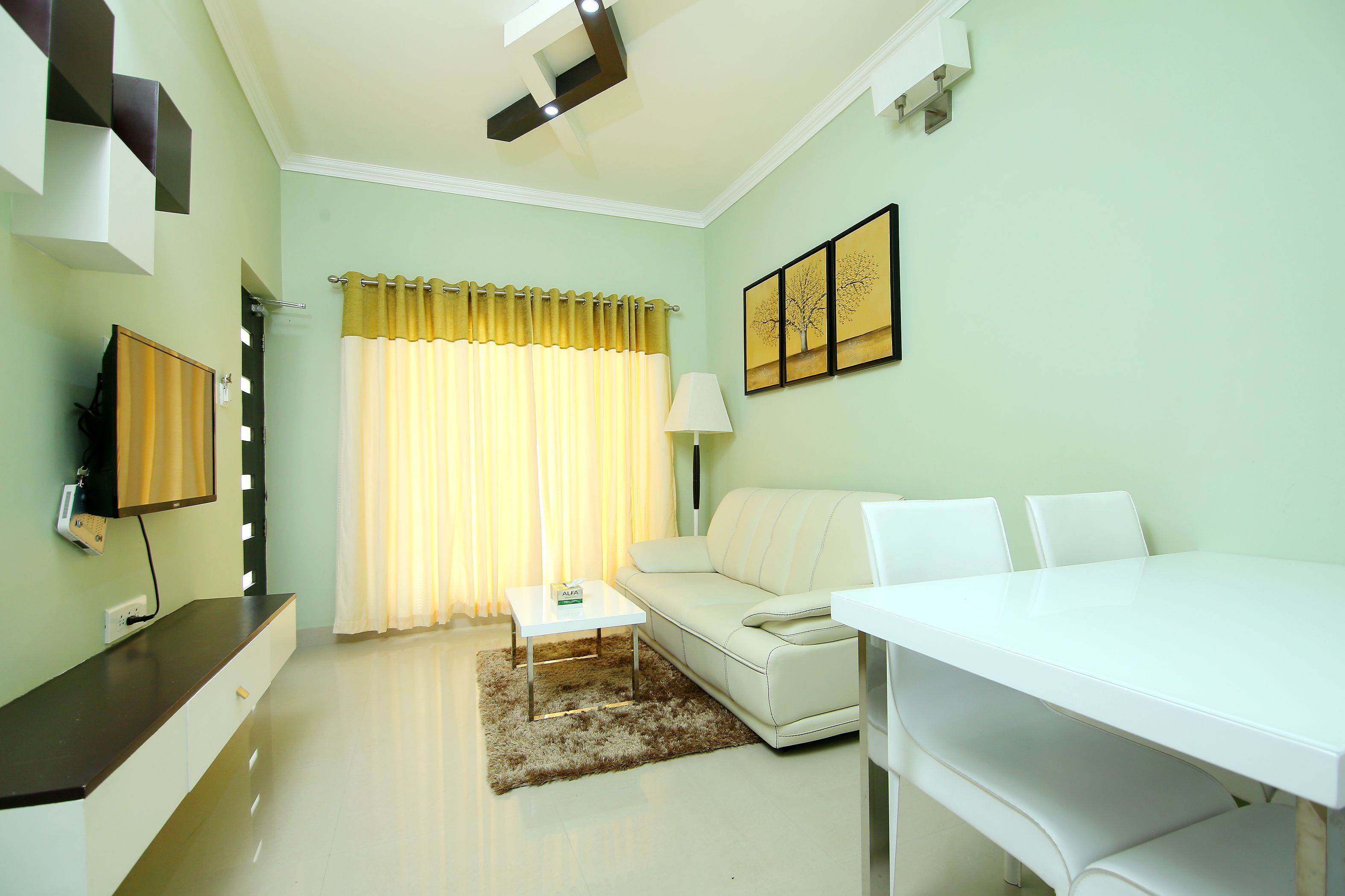 A modern living room with a white sofa, a white dining table, and a flat-screen TV mounted on the wall.