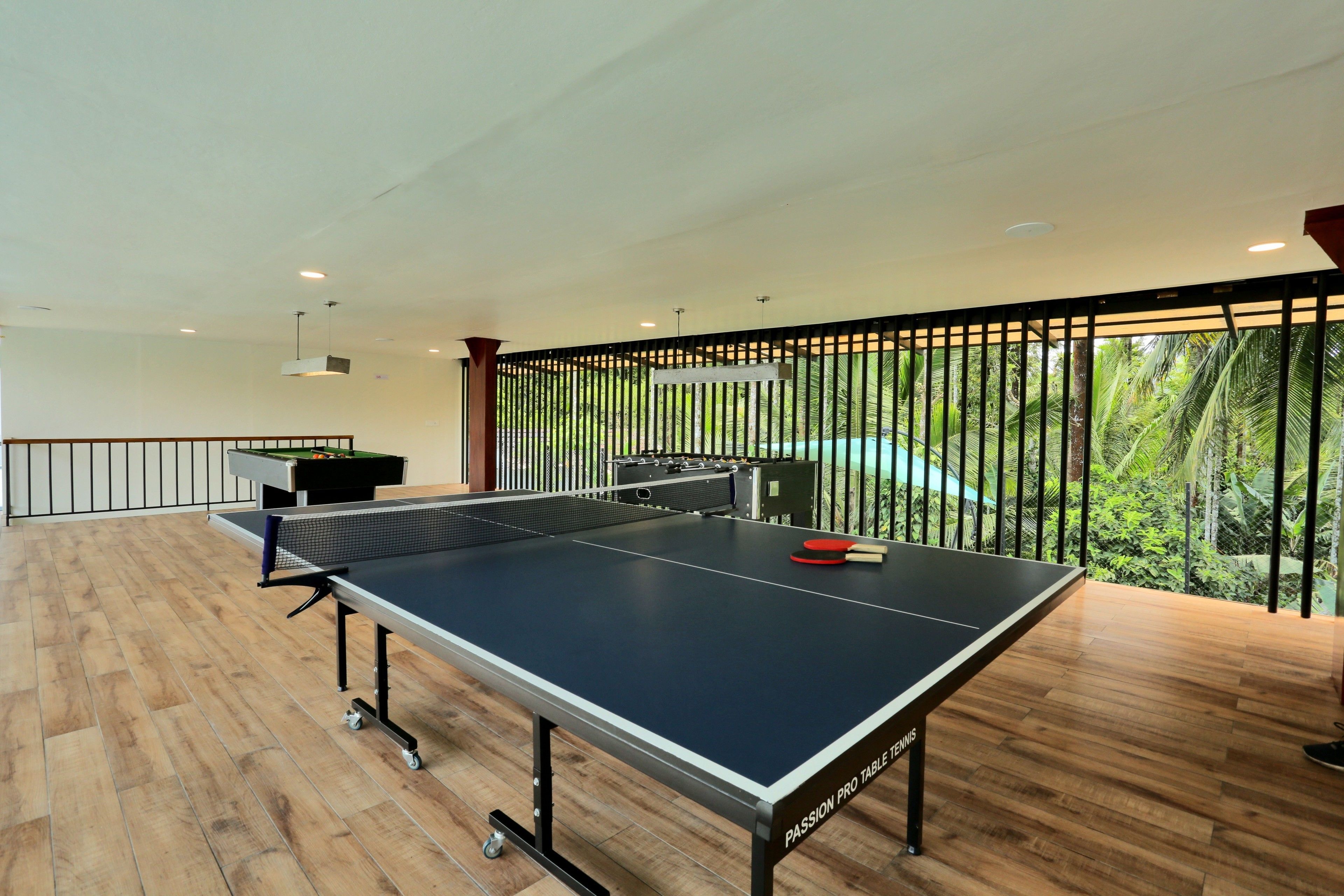A ping pong table in a room with wooden flooring, a pool table, and a view of greenery through a railing.