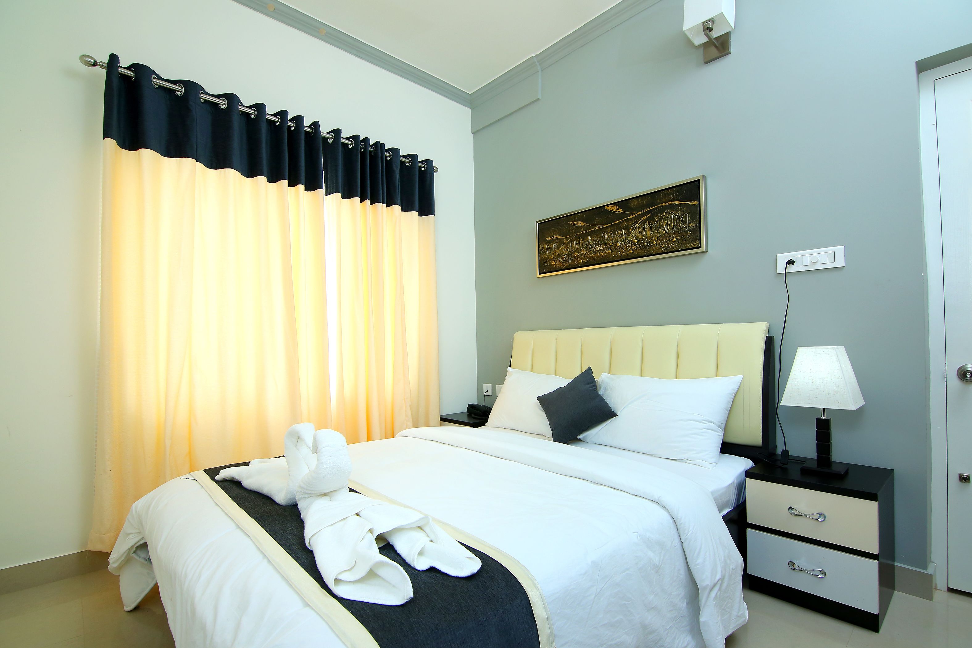 A neatly made bed with white linens and a black throw, a bedside table with a lamp, and a painting on the wall.