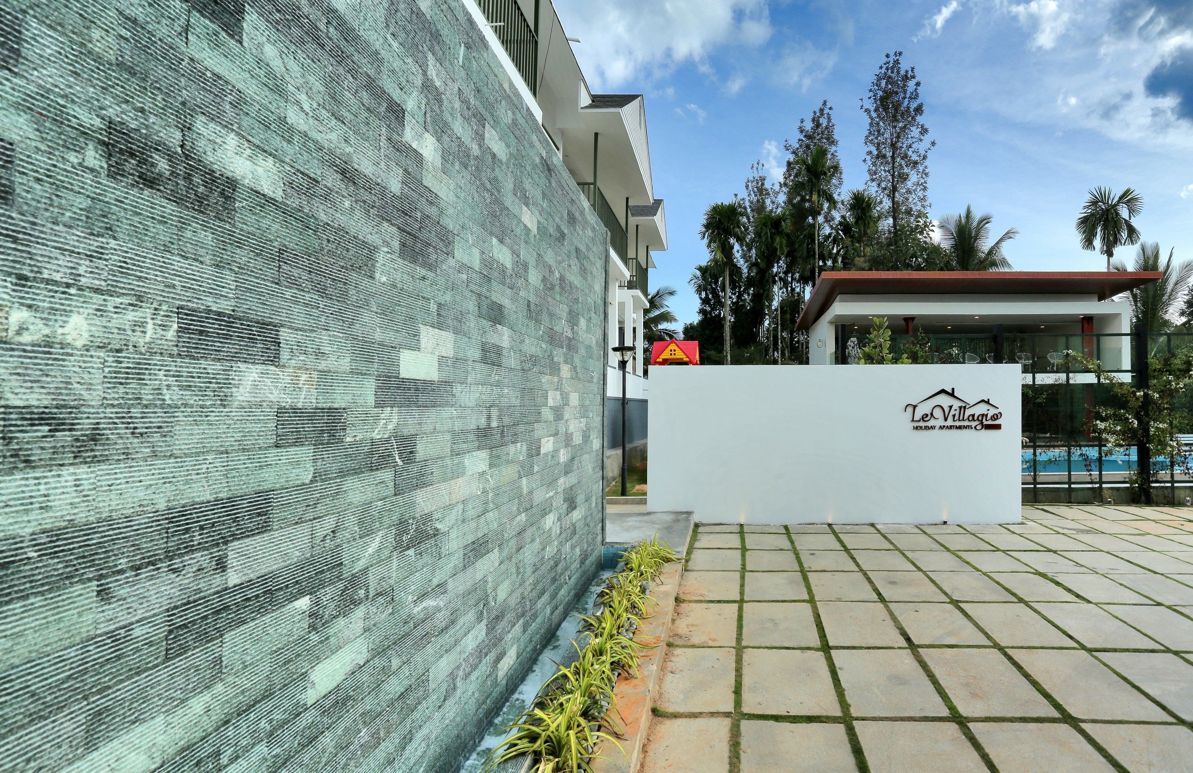 A modern building with a textured facade, a sign reading "Le Village Holiday Apartments," and a pool area with palm trees.