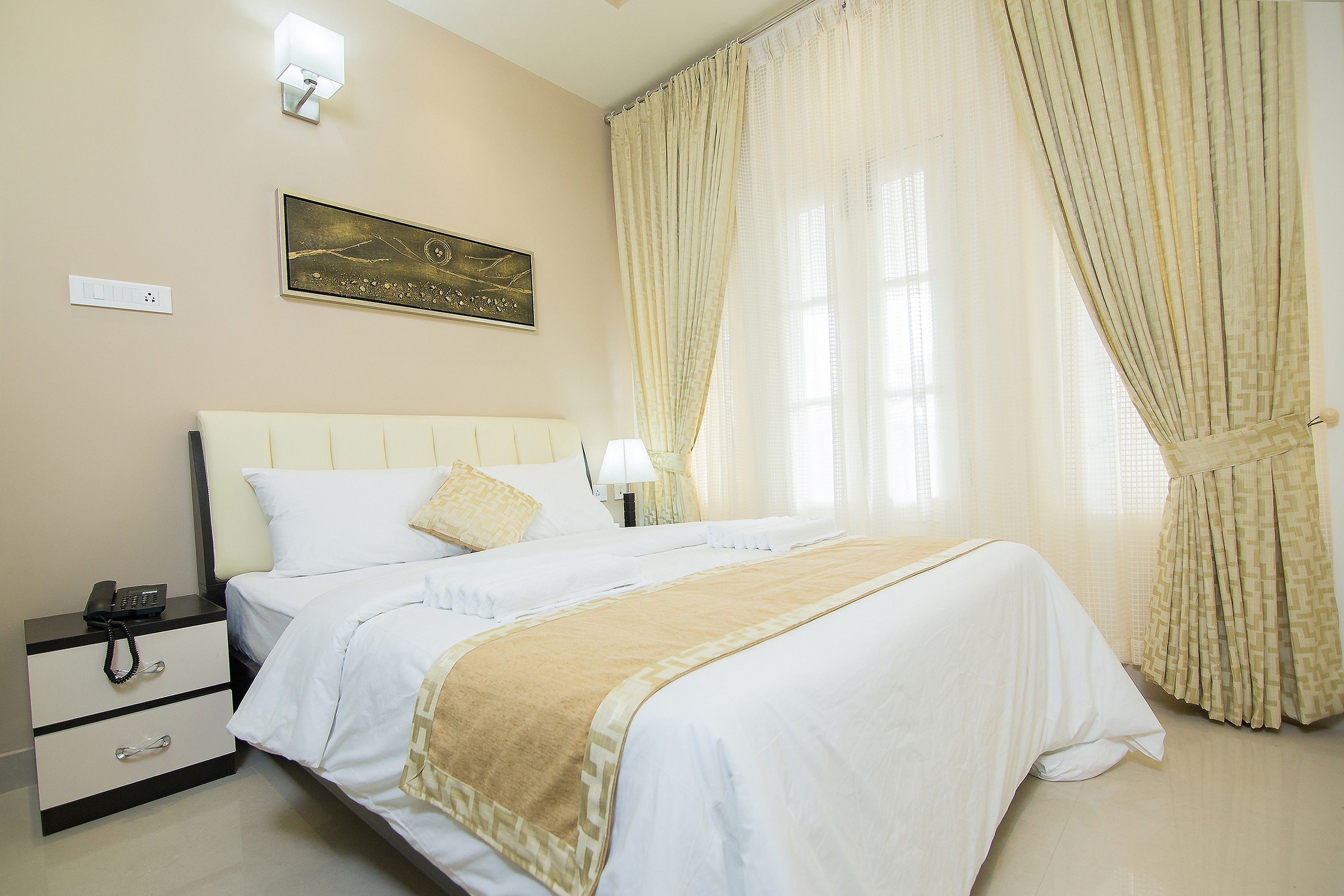 A neatly made bed with a beige throw blanket, a bedside table with a lamp, and a wall-mounted painting.