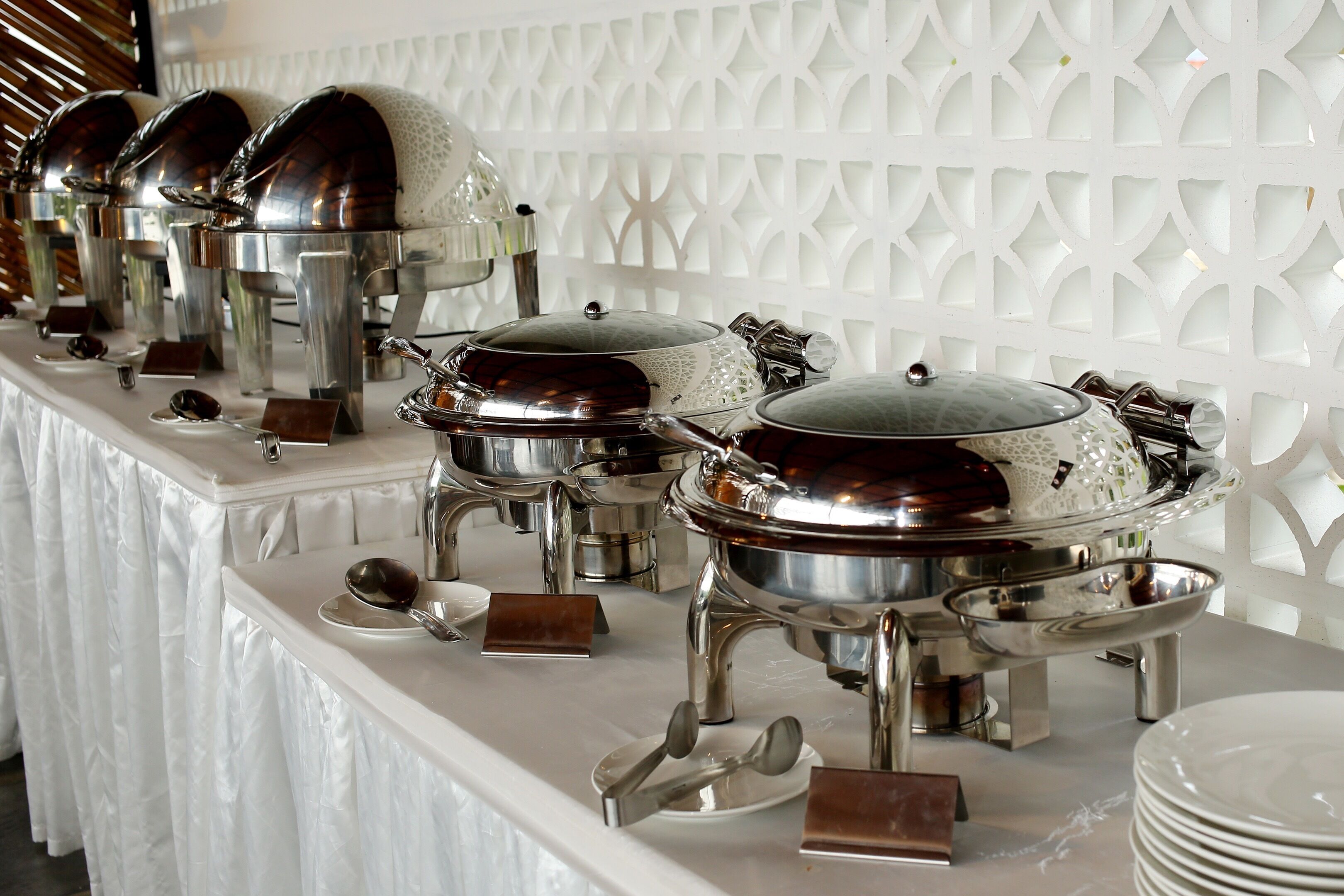 A buffet setup with multiple stainless steel chafing dishes and serving utensils on a white table against a decorative white wall.