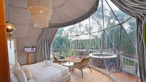 A spacious room with a large bed, a small table, and a chair. There is a TV on a stand and a large window offering a view of the outdoors.