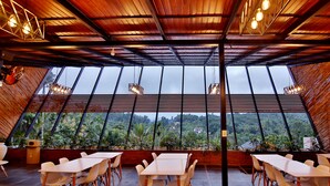 A restaurant with a high ceiling and large windows offering a view of the surrounding greenery. Tables and chairs are arranged for dining.