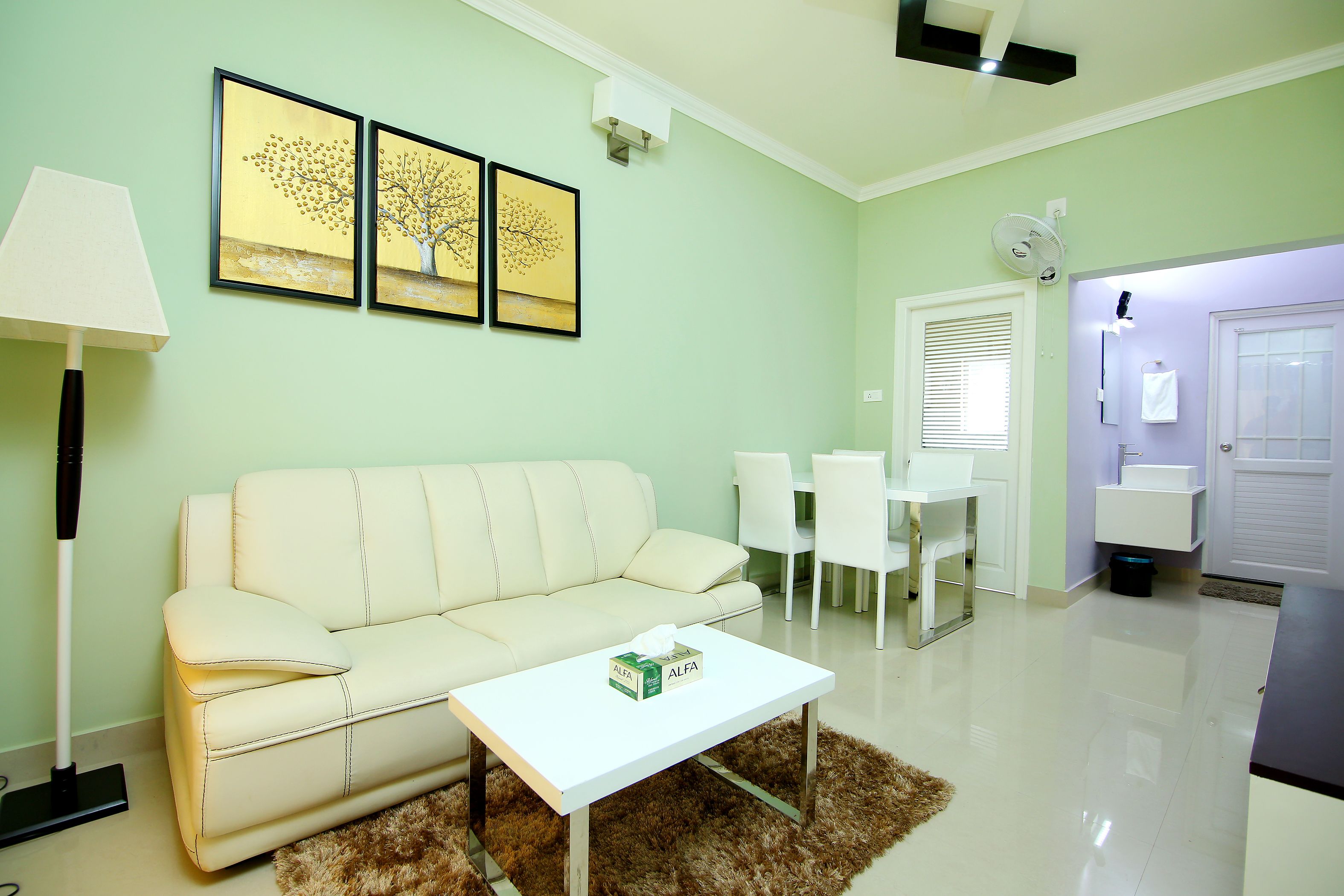 A modern living room with a white sofa, a coffee table, and a dining area with white chairs. There are framed artworks on the wall and a small table with a box on it.