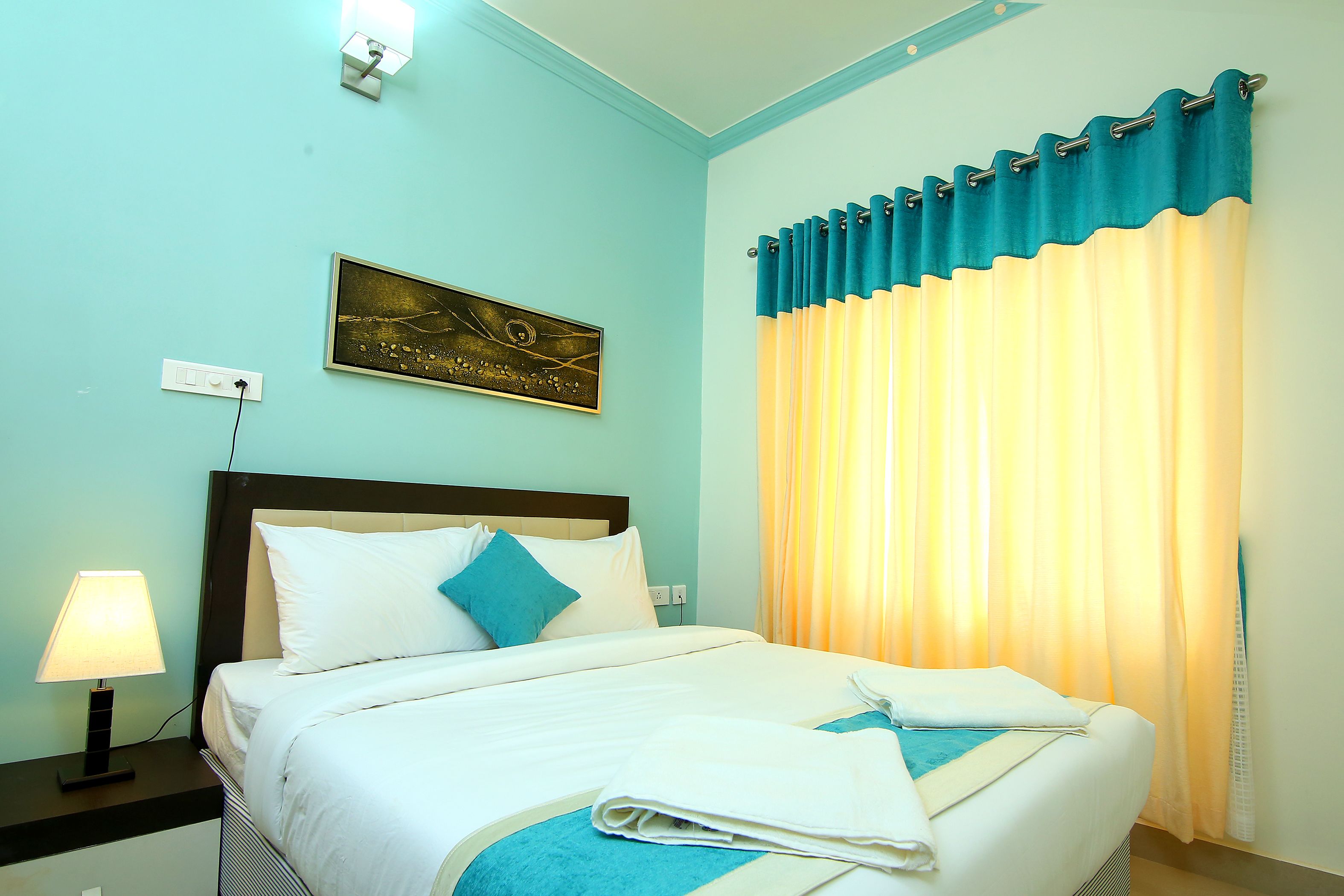 A neatly made bed with white and blue bedding, a bedside lamp, and a wall-mounted painting.