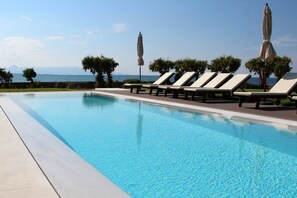 En pool med solstolar och parasoller, en klar himmel och utsikt över havet.