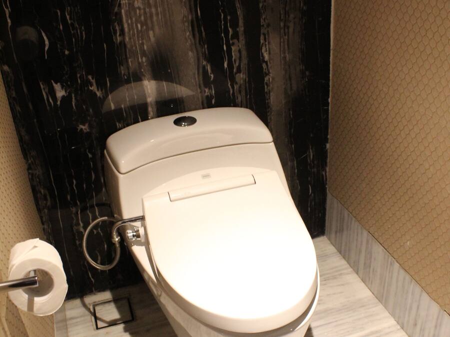 Un inodoro con asiento y tapa blancos, un cubo de basura negro y un suelo de mármol.