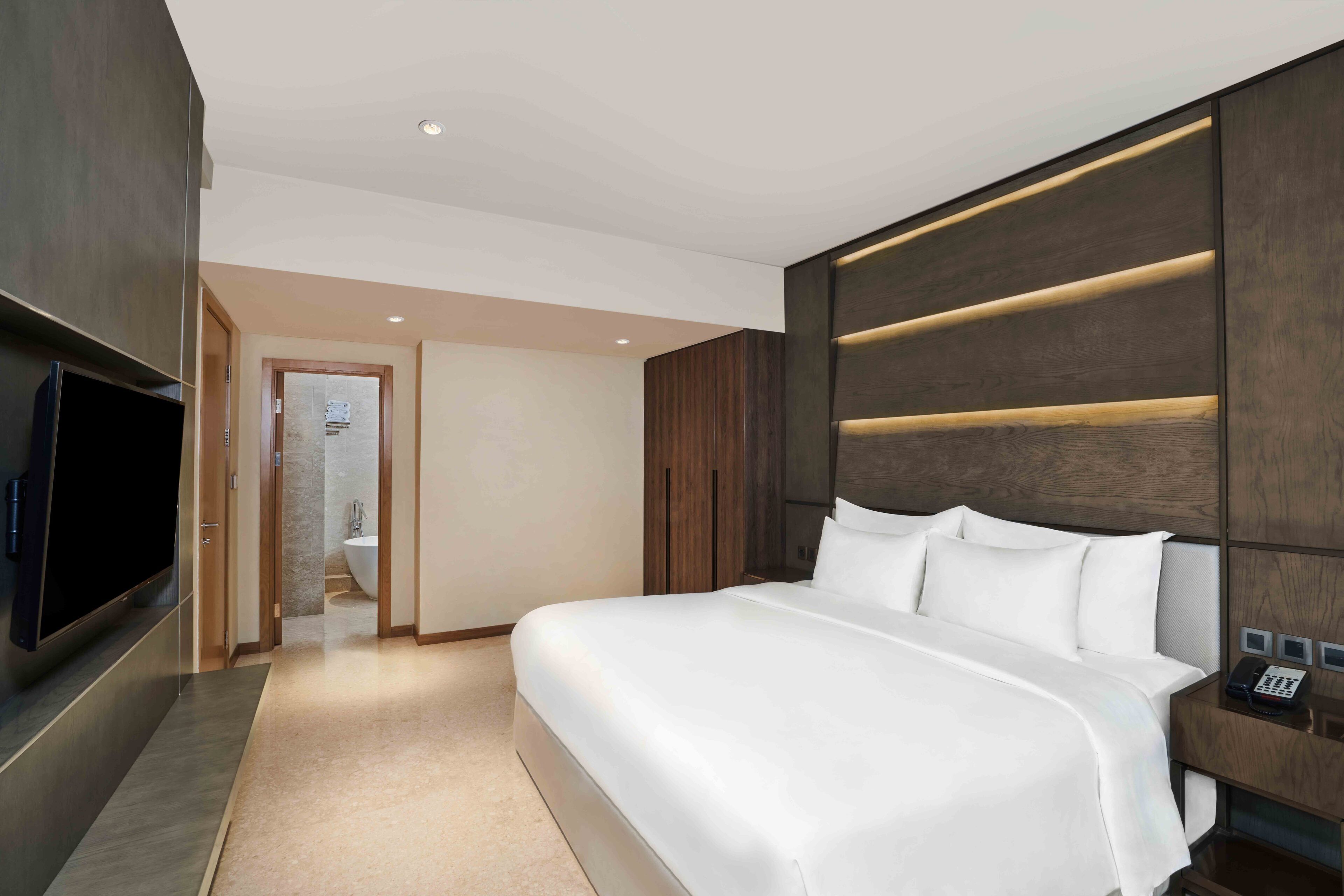 A modern hotel room with a large bed, a flat-screen TV, a bedside telephone, and a bathroom visible through an open door.