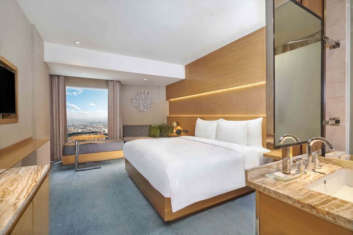 Ett modernt hotellrum med en stor säng, ett badrum med spegel och handfat samt utsikt över staden genom fönstret.