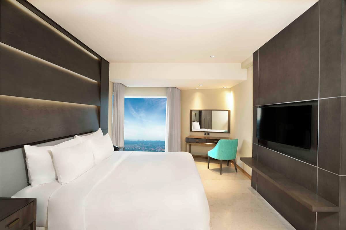 Ett hotellrum med en stor säng, ett skrivbord, en stol, en tv och utsikt över staden.