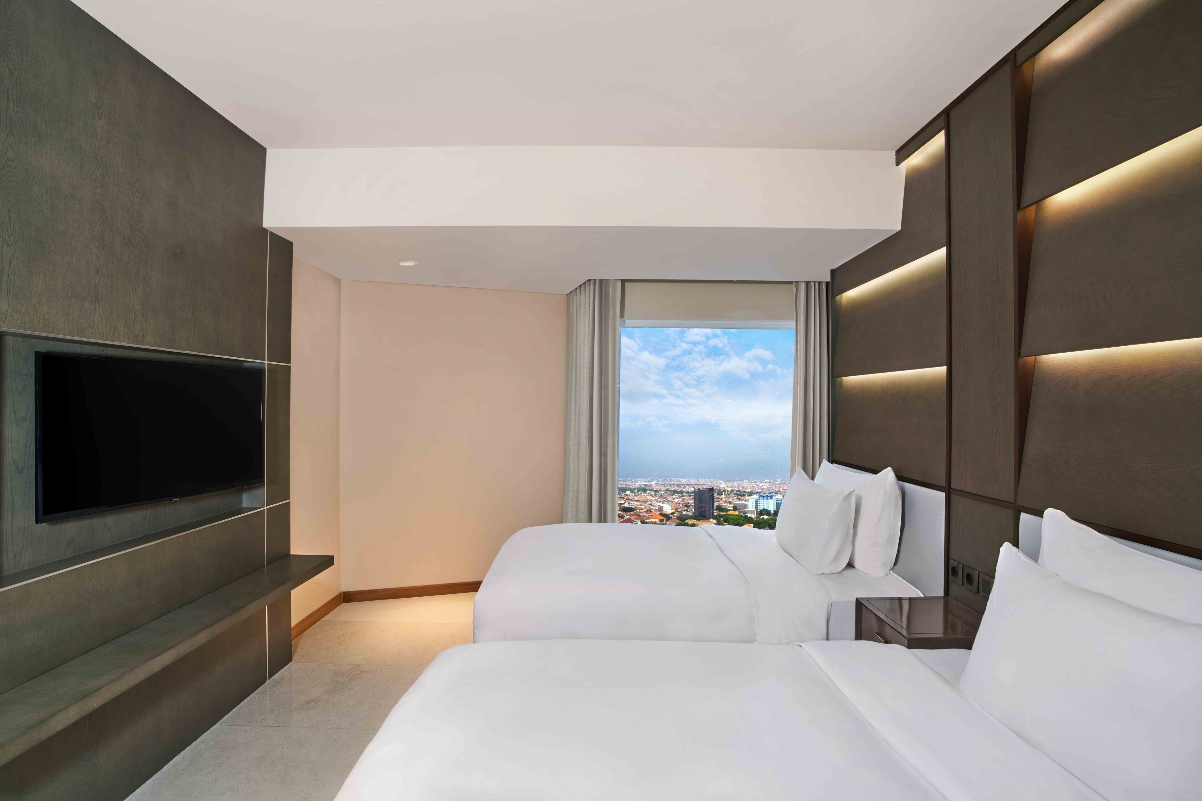 Una habitación de hotel moderna con un televisor grande, dos camas, vistas a la ciudad y un diseño minimalista.