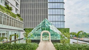 Un edificio moderno con una estructura piramidal de vidrio en el centro, rodeado de vegetación y un sendero que conduce hacia él.