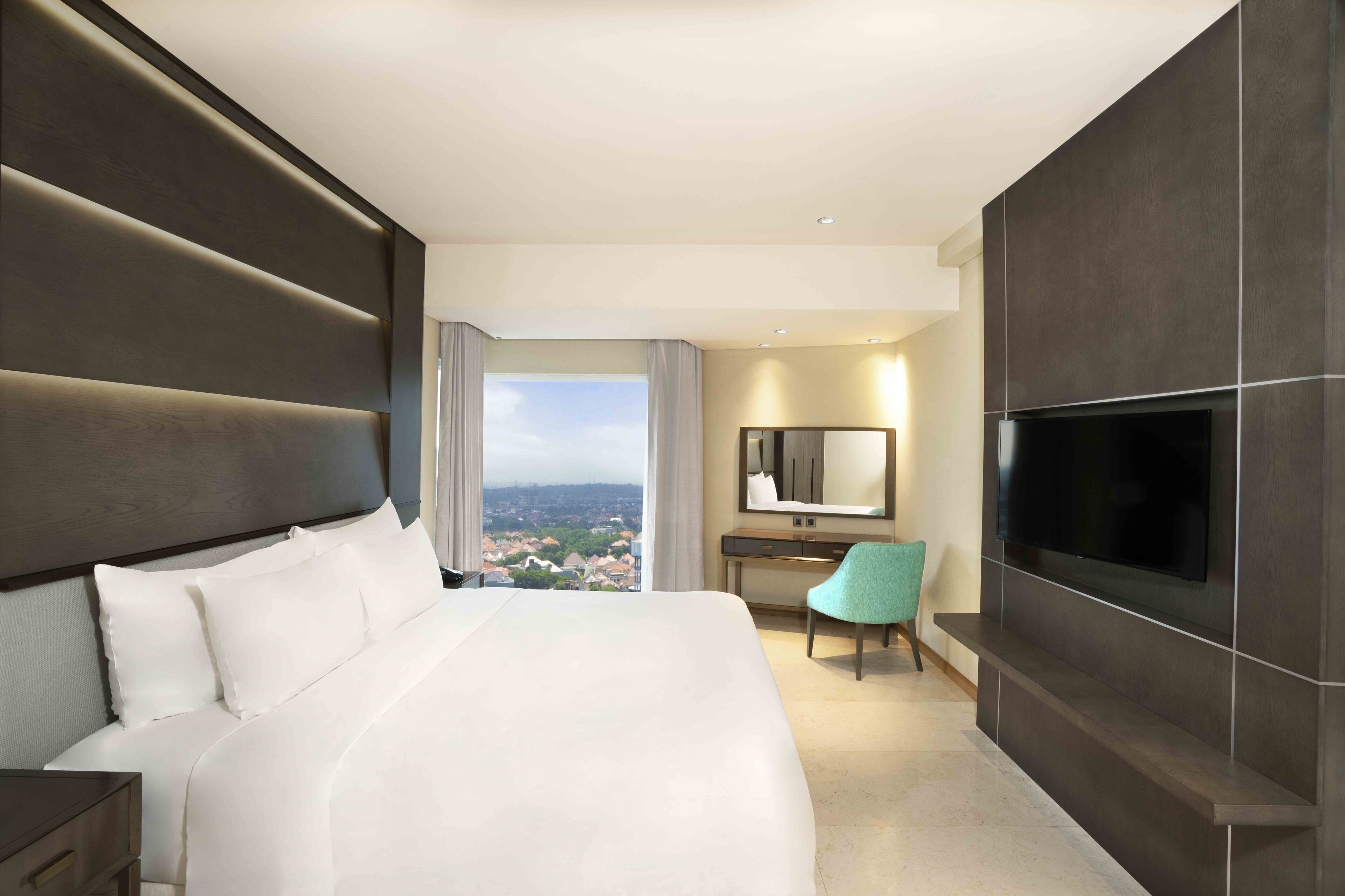 A modern hotel room with a large bed, a desk, a chair, a TV, and a city view.
