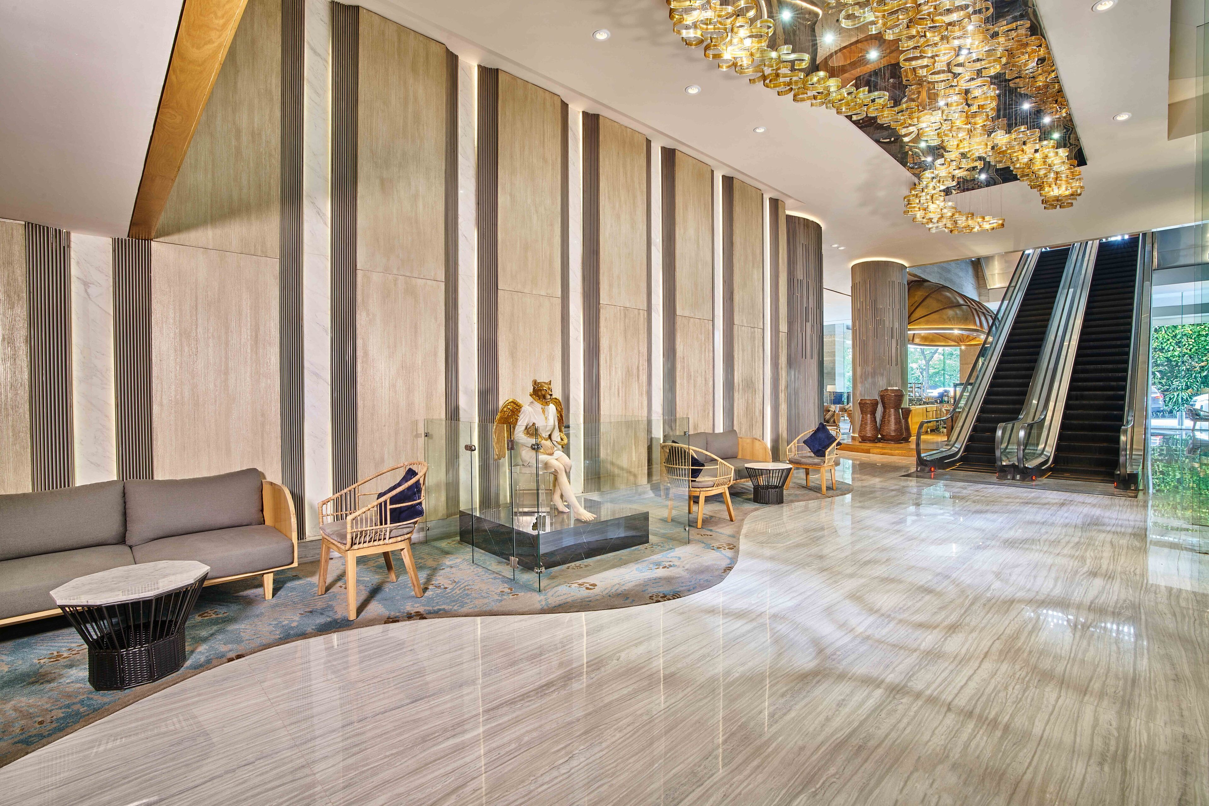 A spacious lobby with a modern design, featuring a seating area with a sofa and chairs, a glass display case with a statue, and an escalator.