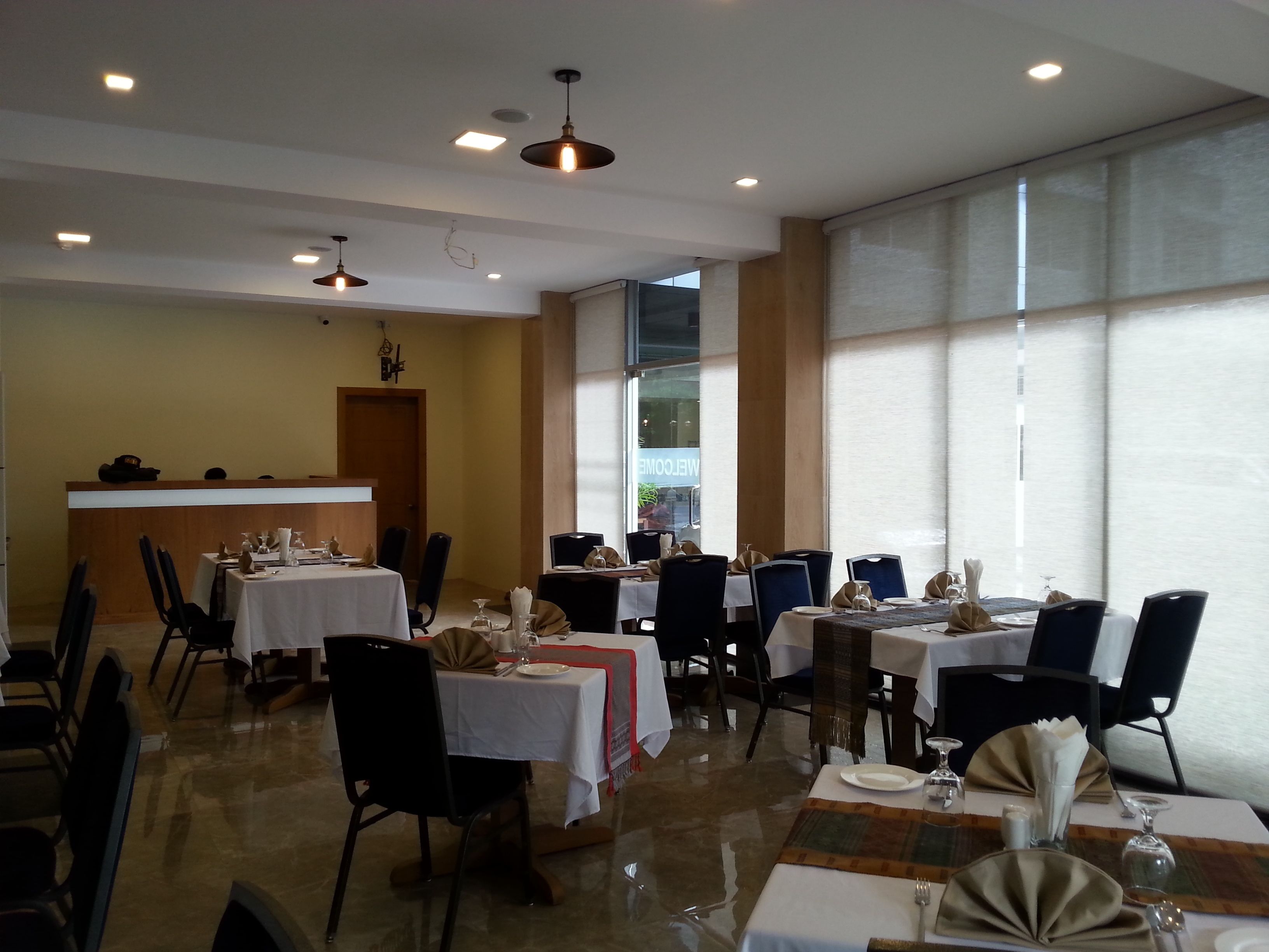 A dining area with multiple tables and chairs, a reception desk, and large windows allowing natural light.