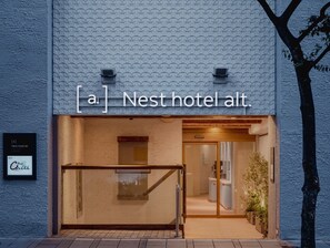 L’extérieur de l’hôtel Nest Alt. se distingue par sa façade texturée, une entrée bien visible et une enseigne indiquant la présence d’un bar et d’un espace de détente.