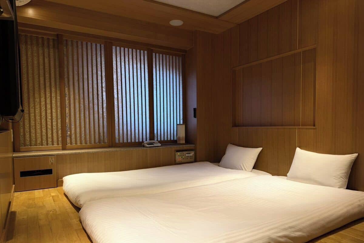 A hotel room with a large bed, wooden paneling, a window with blinds, and a bedside table with a lamp.