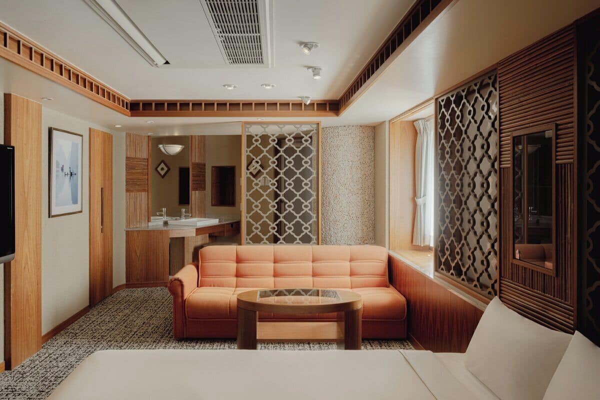 A modern hotel room with a sofa, a bed, a small table, a TV, and a bathroom visible through a partition.