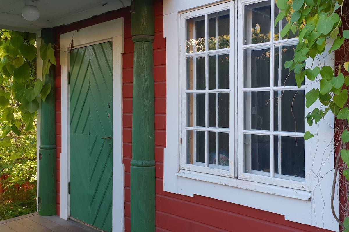 Ein rotes Gebäude mit grüner Tür und grünem Fenster, umgeben von viel Grün.