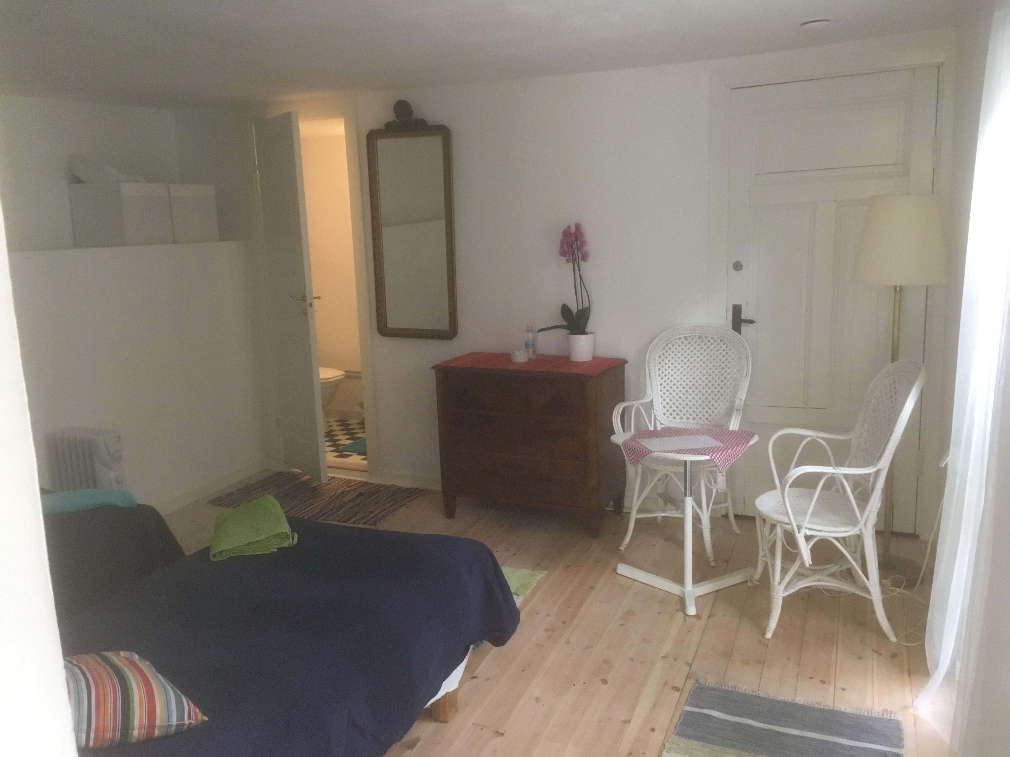 Una camera da letto con un letto, due sedie bianche, una cassettiera, uno specchio e una porta.