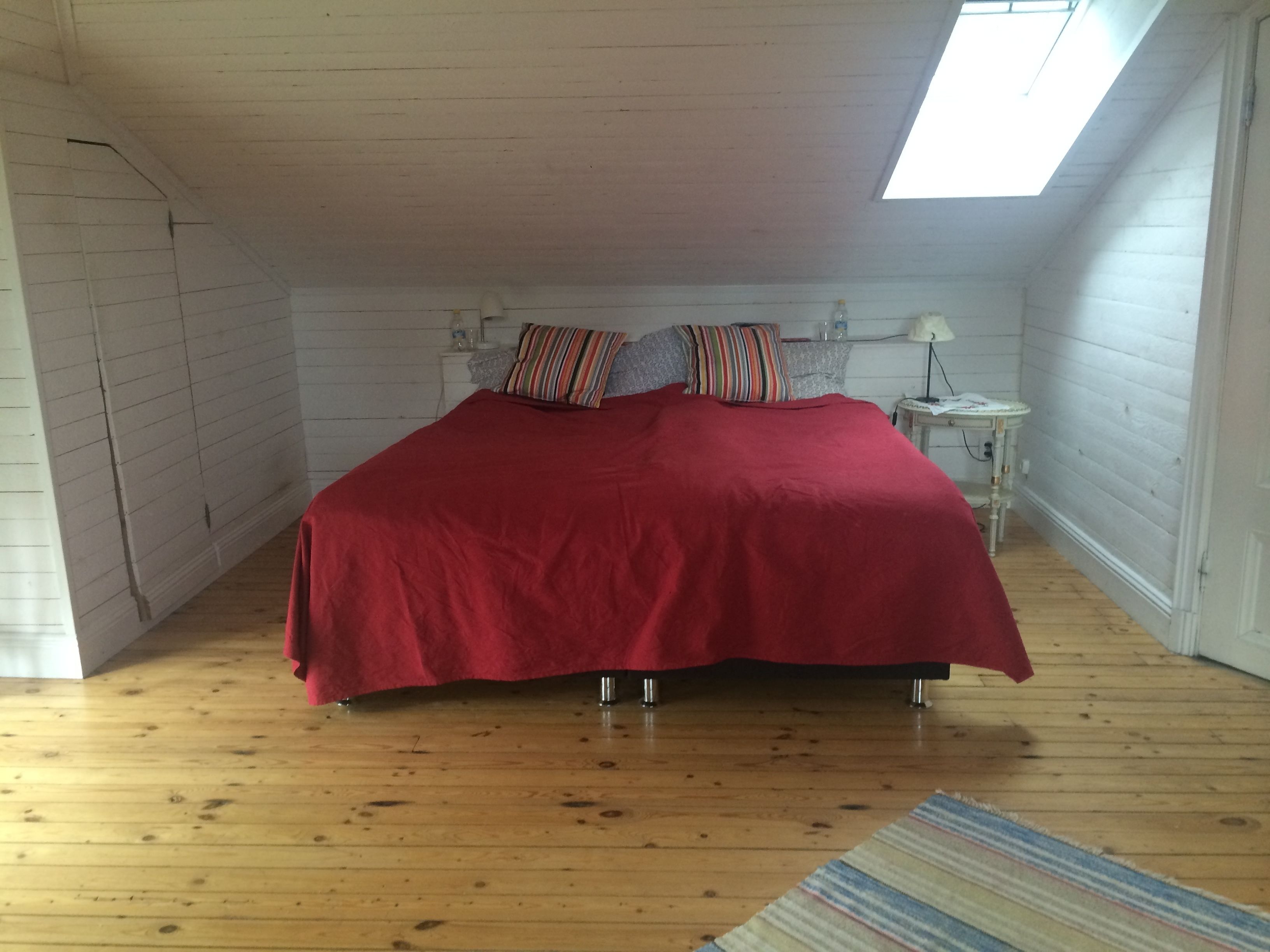 A bedroom with a red bedspread, a skylight, wooden walls, a small bedside table, and a nightstand.