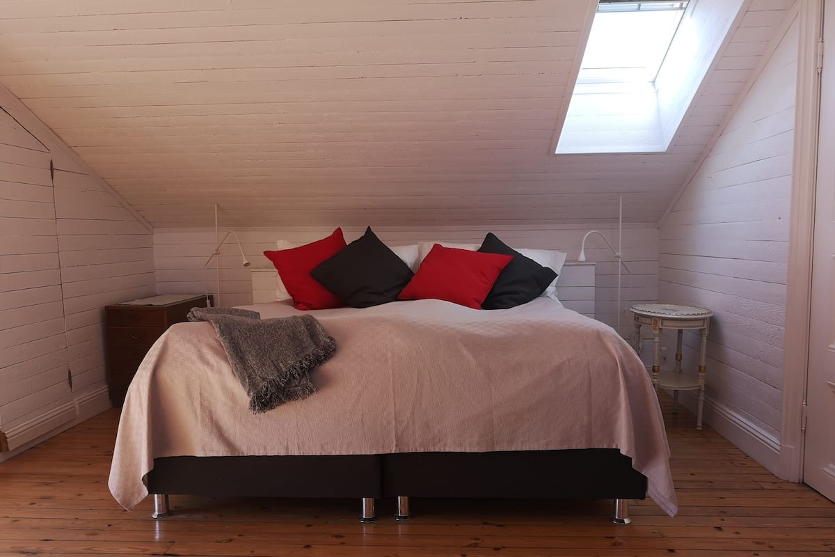 Ein Schlafzimmer mit schrägverdeckter Decke, einem Bett mit beiger Bettdecke und roten und schwarzen Kissen, einem kleinen Nachttisch und einem Fenster mit Oberlicht.