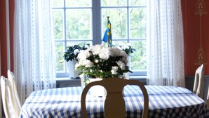 Una mesa de comedor con mantel a cuadros, rodeada de sillas blancas. En la mesa, un jarrón con flores blancas y una bandera.