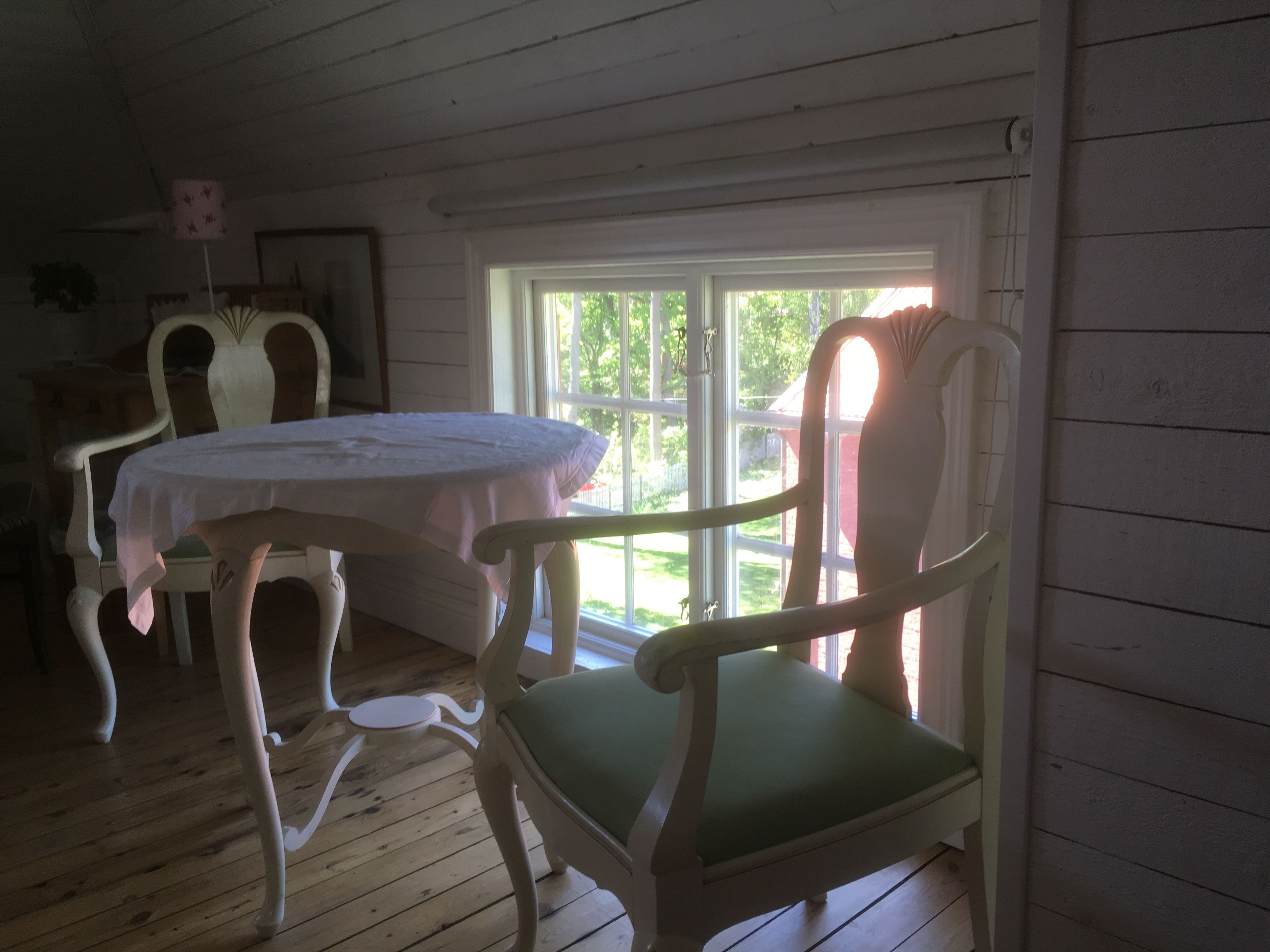 Een kamer met een houten vloer, een ronde tafel met een witte tafelkleed en twee stoelen met groene kussens.