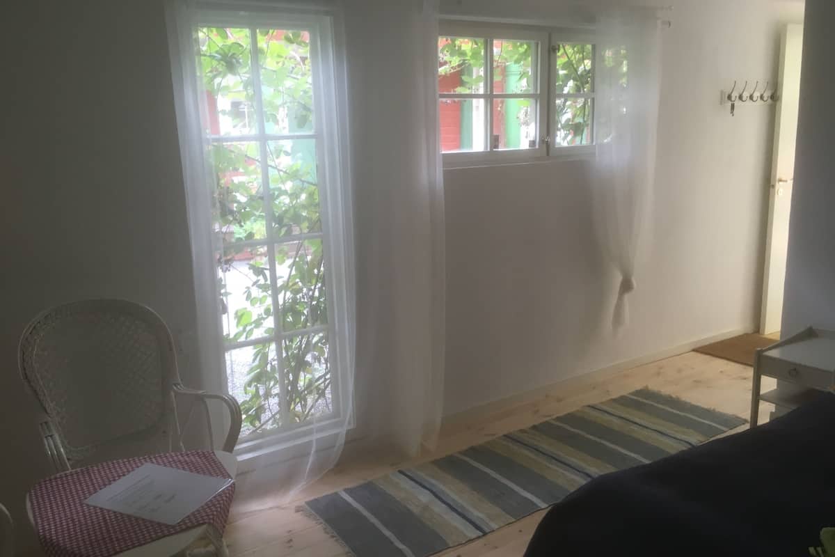 Ein Zimmer mit Holzboden, einem Streifenmuster-Teppich, einem Stuhl mit karierter Tischdecke und einem Fenster mit Blick ins Grüne.